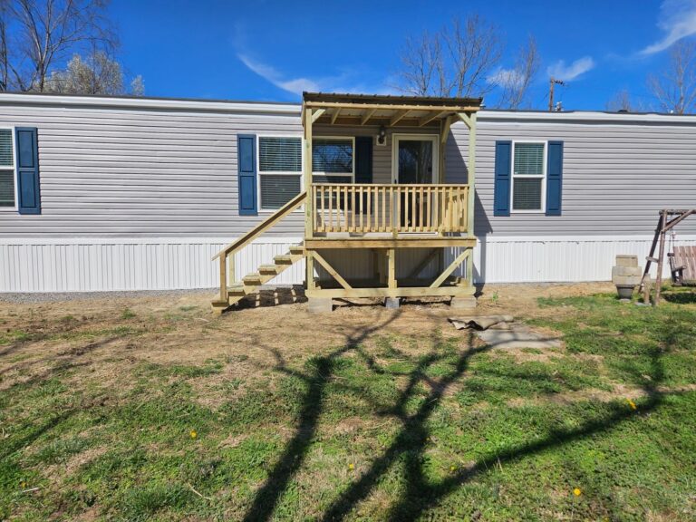 Angled view of a 6x10 covered front deck with a wooden staircase and railing attached to a light gray mobile home in Elkton, Kentucky.