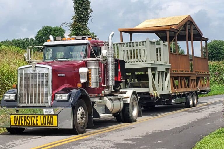 Portable pavilion and deck on the back of a truck with oversize load sign. Truck is on road next to a cornfield