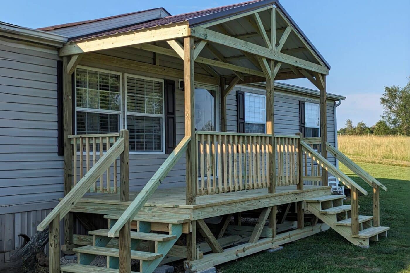 Close up of mobile home porch attached to home near field with steps, railing, and view of foundation