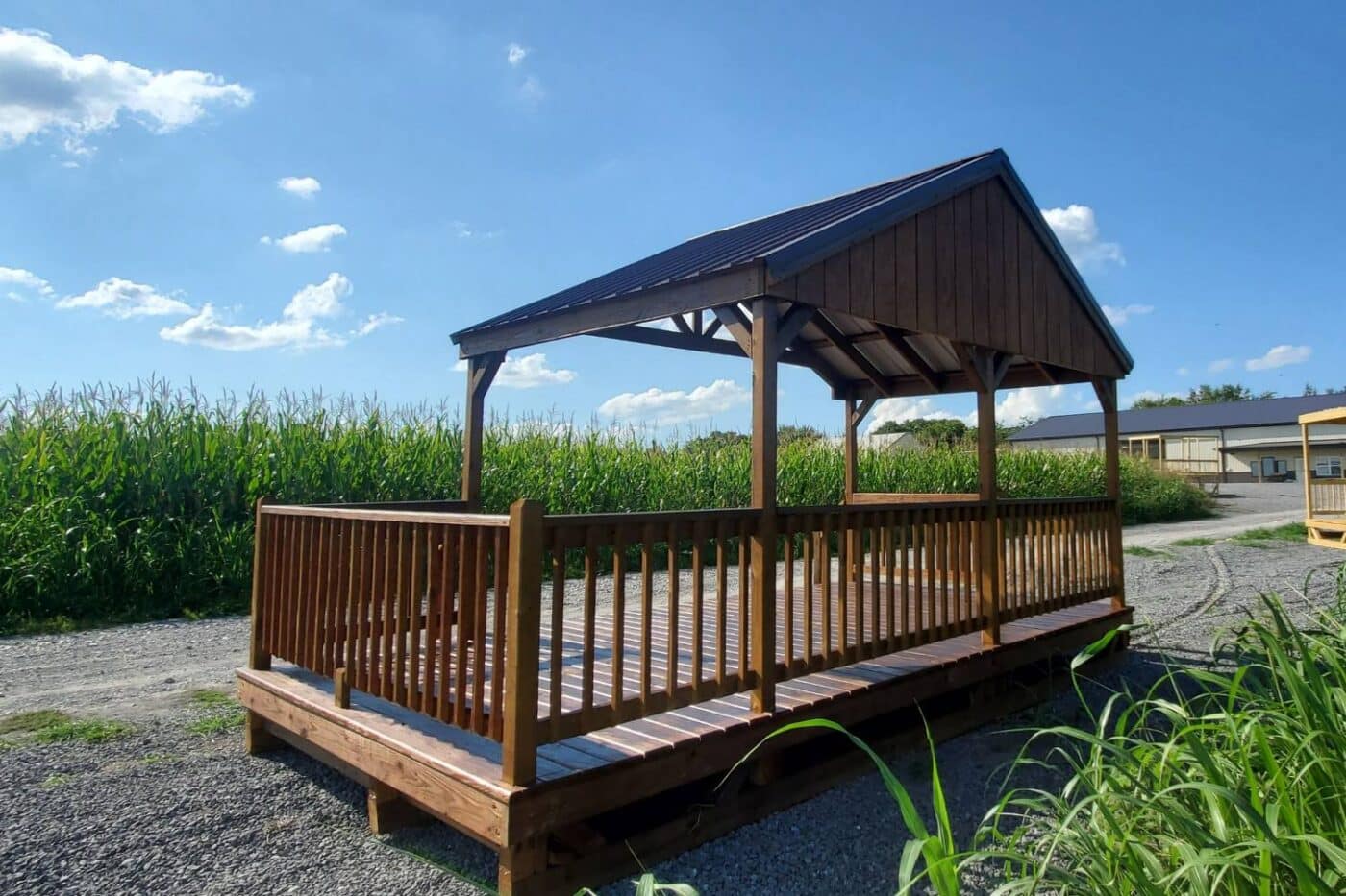 Pavilion on gravel road besides cornfield. Pavilion is stained dark wood with additional deck area