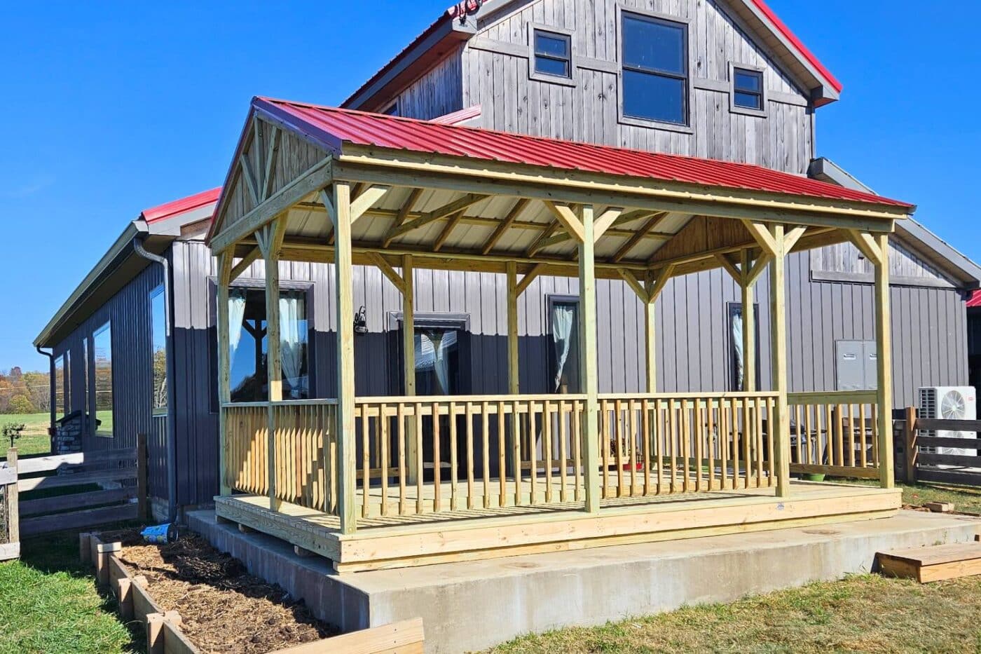 Wooden pavilion besides barn with red metal roof and wooden railing. Pavilion sits on large concrete block