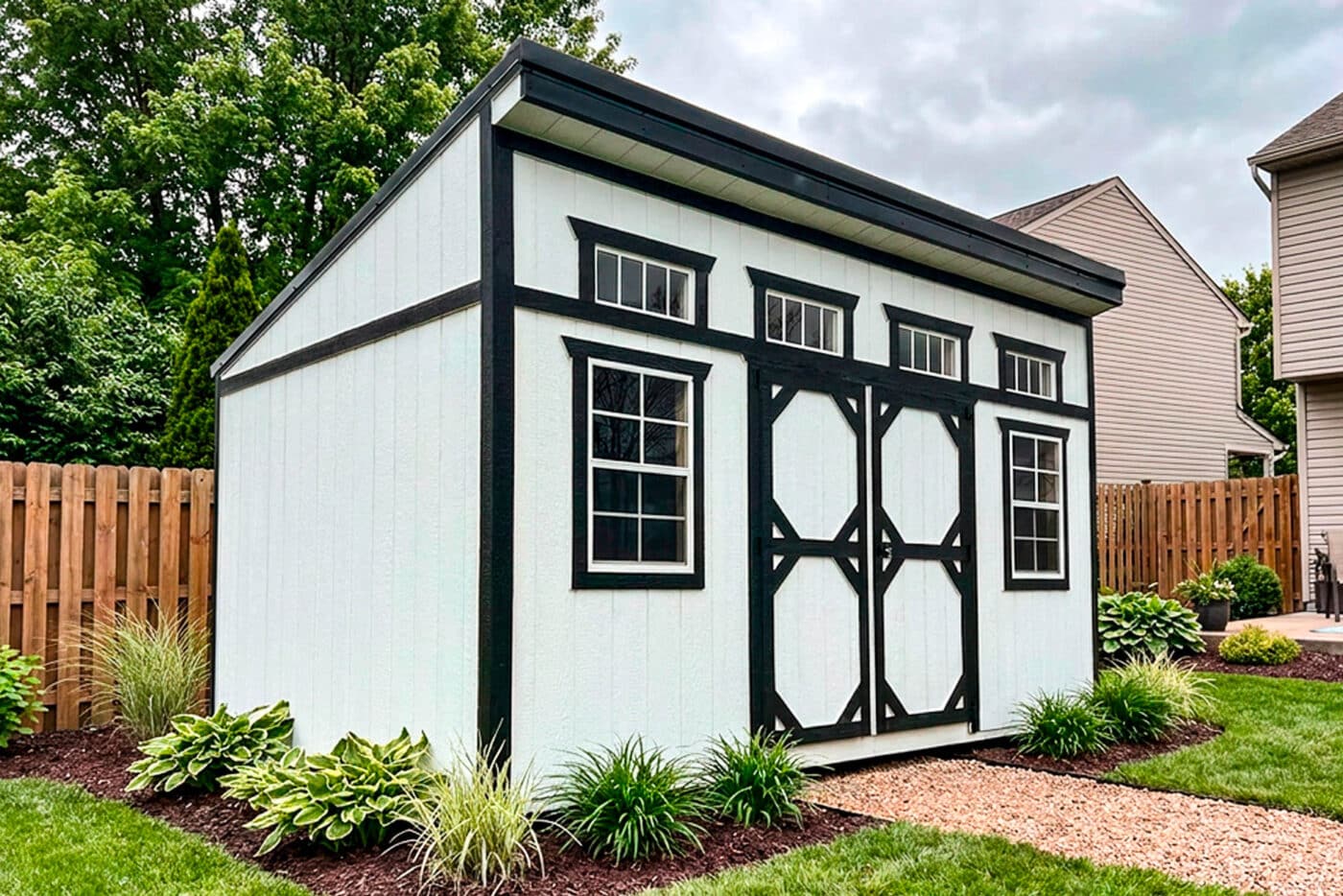 White modern shed with black trim, double doors, two windows, and four transom windows beside a landscaped yard.