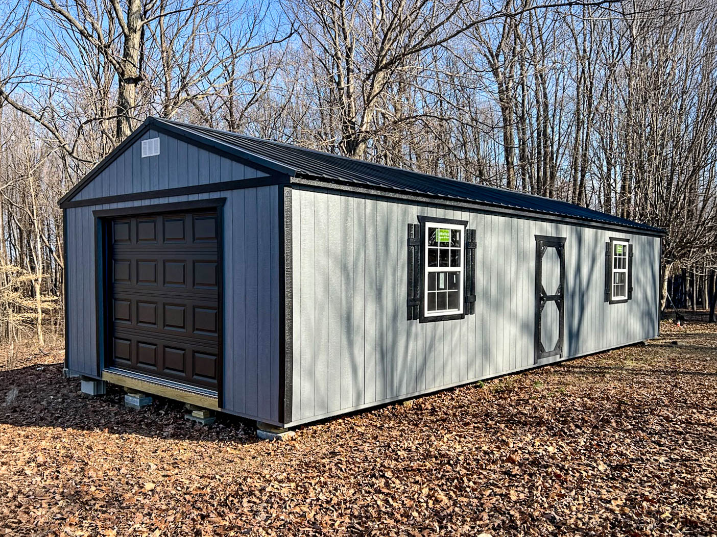 Gray garage with black trim, overhead door, side entry door, two windows, and a black metal roof.