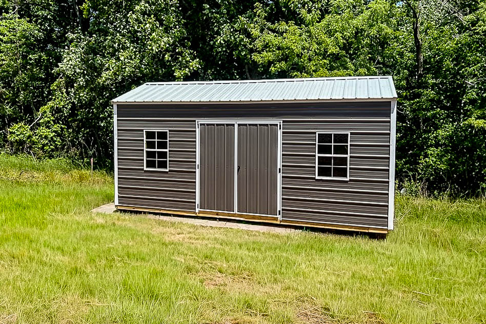Gray metal storage shed with double doors and two windows in a grassy yard.
