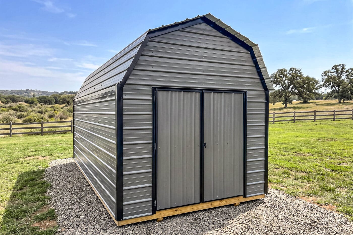 Gray horizontal metal shed with black trim and double doors on a gravel pad.